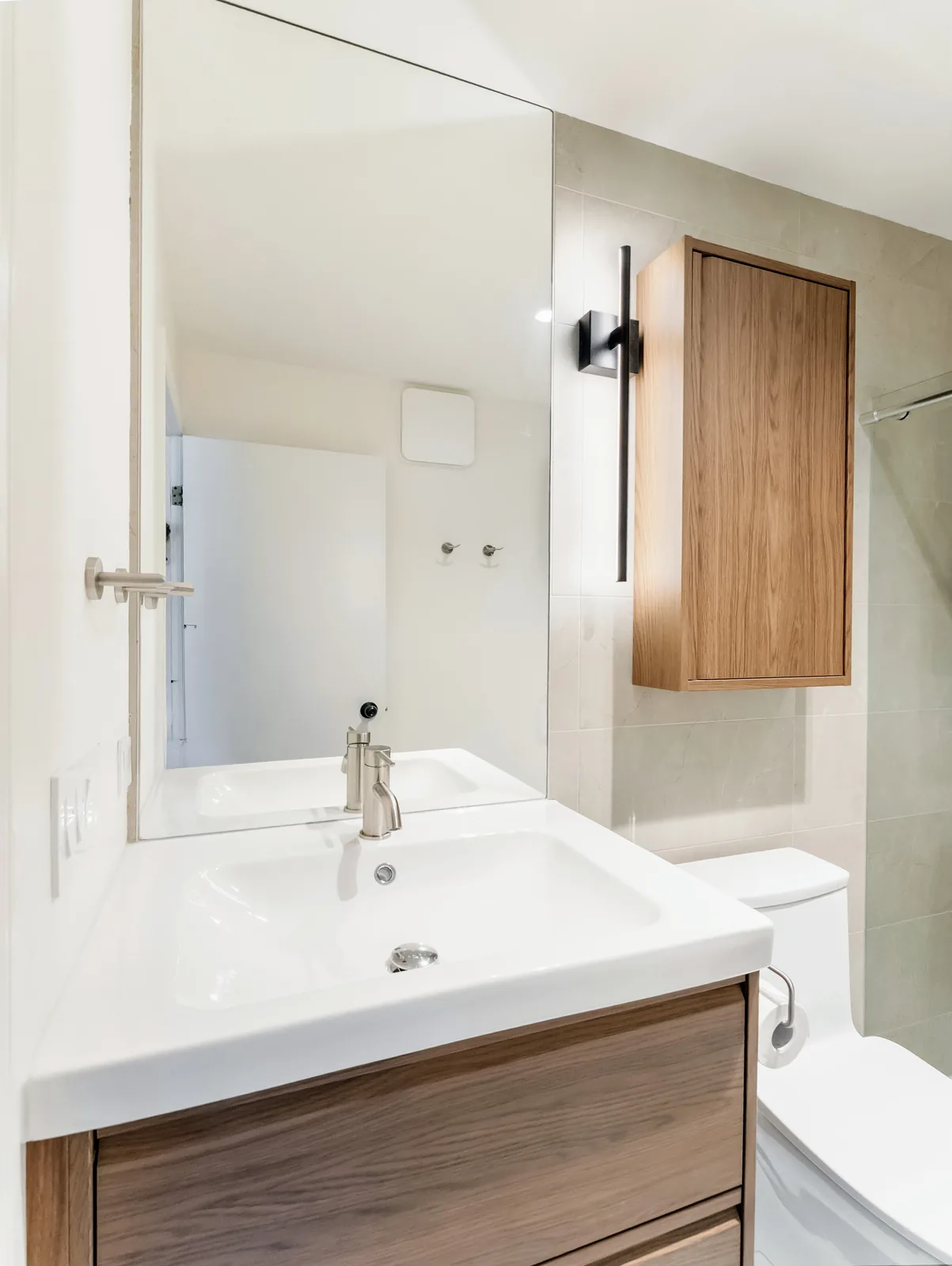 Vanity under a full-width mirror, with a slim black sconce beside the oak wall cabinet