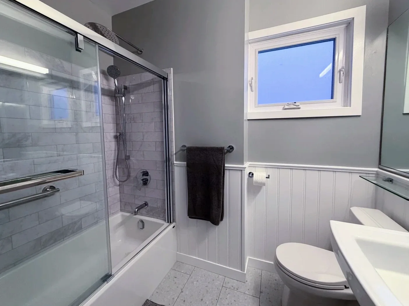 Bathroom in white beadboard under grey walls, with a marble-look tub surround behind sliding glass
